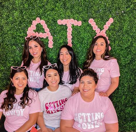 Group photo of six smiling women in matching pink 'Getting' T‑shirts and playful sunglasses, posing in front of a green boxwood wall decorated with pink rose letters for a festive party backdrop.
