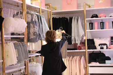 Shopper browsing a sparkly dress on a gold clothing rack in a pink-and-gold women's boutique with denim jackets, hoodies, skirts and accessory shelves