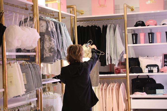 Shopper browsing a sparkly dress on a gold clothing rack in a pink-and-gold women's boutique with denim jackets, hoodies, skirts and accessory shelves