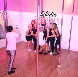 Group of women in black activewear and colorful pink and purple wigs posing for a fun photo in a pink-lit pole-fitness dance studio with vertical poles, someone capturing it on a smartphone.