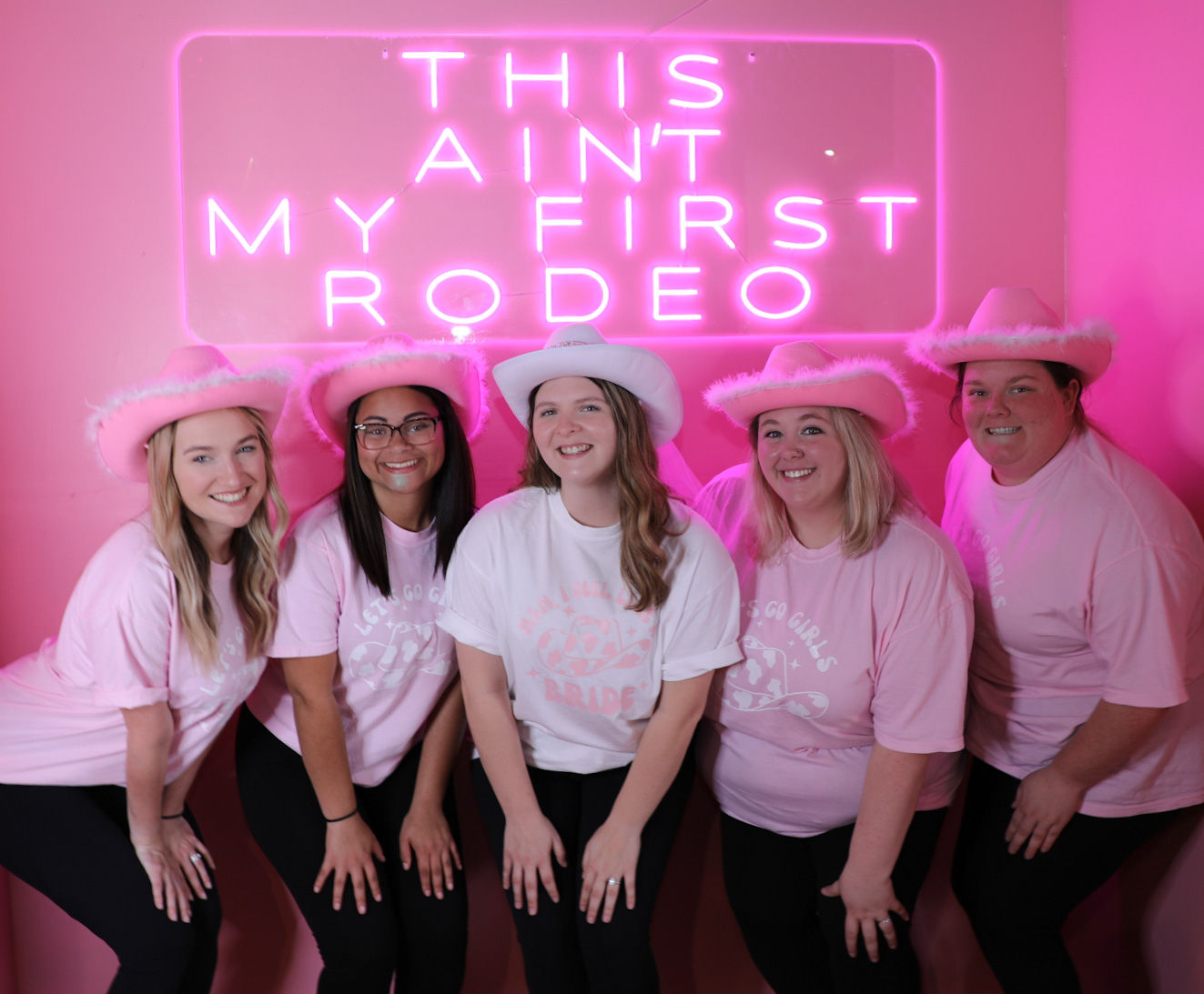 Five women in pink fuzzy cowgirl hats and matching shirts smiling in front of a bright pink neon sign reading THIS AIN'T MY FIRST RODEO, playful bachelorette party photo.