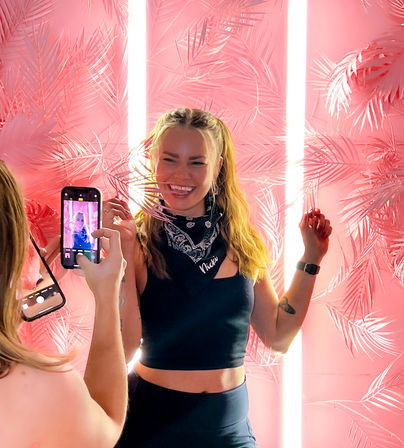 Smiling woman in a black crop top and bandana posing for a phone photo in front of an Instagrammable neon-lit pink palm-leaf backdrop