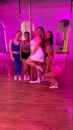 Five women in activewear posing around a chrome pole in a pink neon-lit pole-dance studio with mirrored arches and hardwood floors.