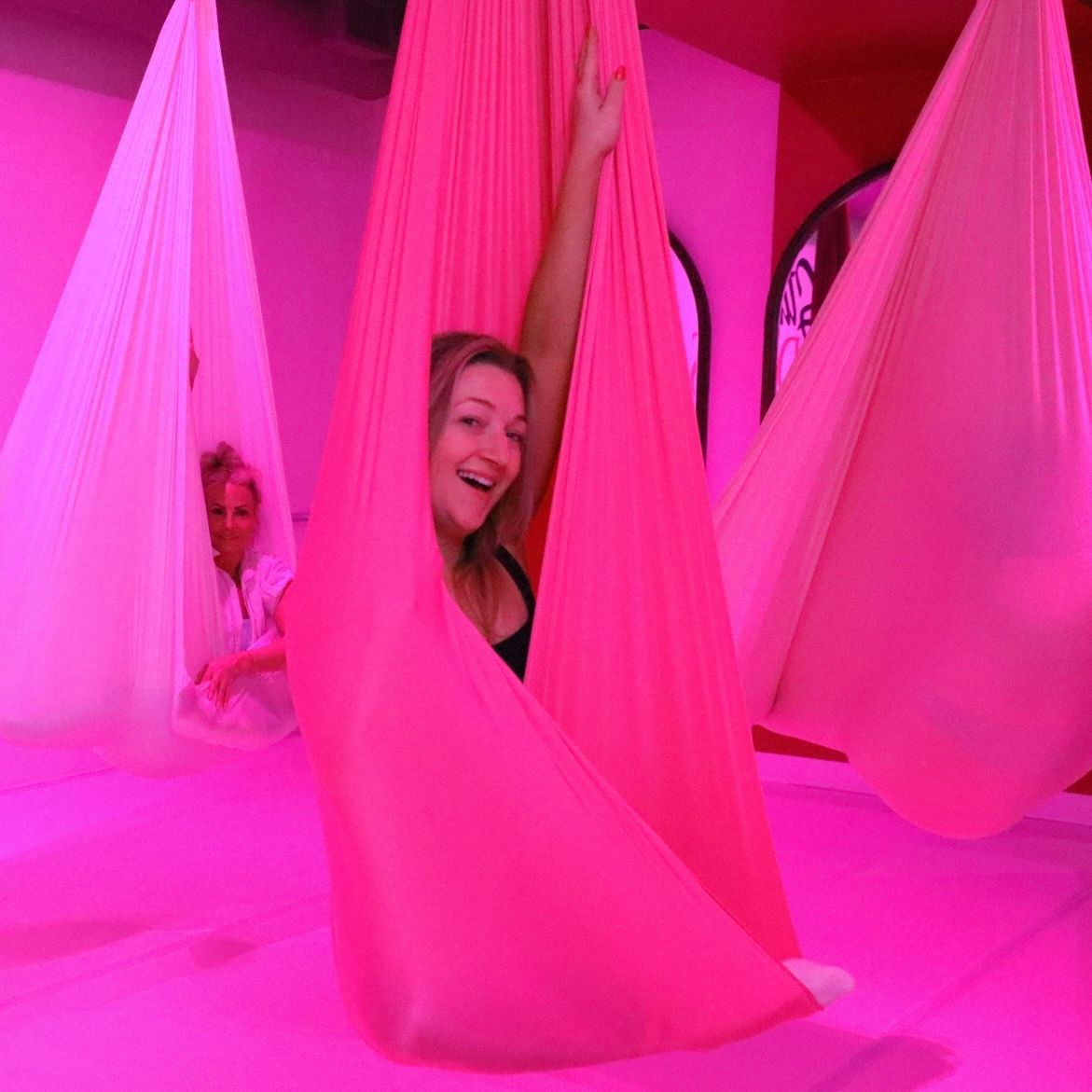Smiling woman peeking out of a bright pink aerial yoga hammock in a neon-pink studio, with another person relaxing in a white hammock in the background.