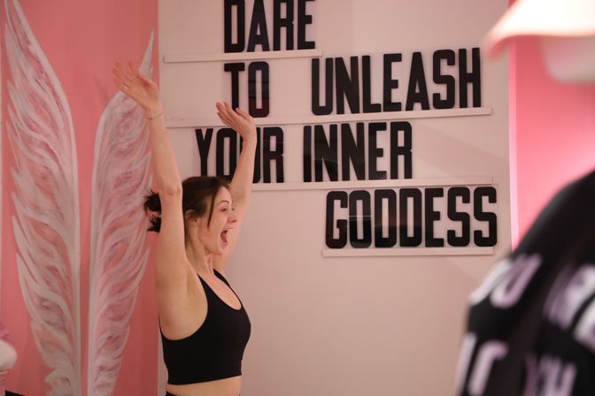 Woman in black sports bra celebrating with arms raised in a pink workout studio, posing in front of painted angel wings and a marquee sign reading 'Dare to unleash your inner goddess'.