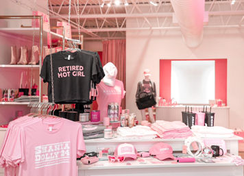 Pink boutique retail display with women's apparel and accessories — racks and a table of graphic tees (one reads “RETIRED HOT GIRL”), pink caps, sunglasses, candles and a mannequin in a bright store interior.