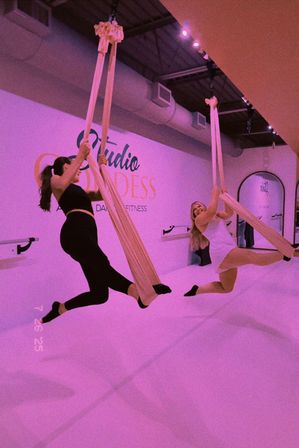 Two people in a pink-lit aerial yoga and fitness studio swinging on suspended silk hammocks, smiling and holding split-leg poses above a padded floor.