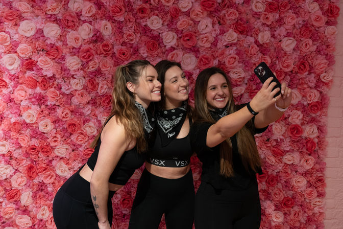 Three smiling friends in black workout clothes and bandanas taking a selfie in front of a bright pink rose wall backdrop.
