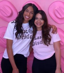 Two smiling friends posing at a bright pink photo wall decorated with pink hats, wearing retro disco-themed graphic tees and black pants