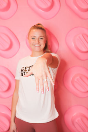 Smiling woman extends her hand to display a sparkling engagement ring in front of a playful pink photo wall with oversized stud shapes