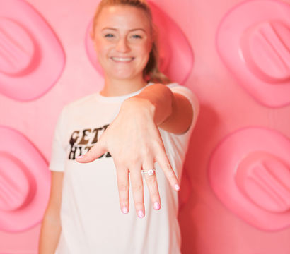 Smiling woman extends her hand to display a sparkling engagement ring in front of a playful pink photo wall with oversized stud shapes