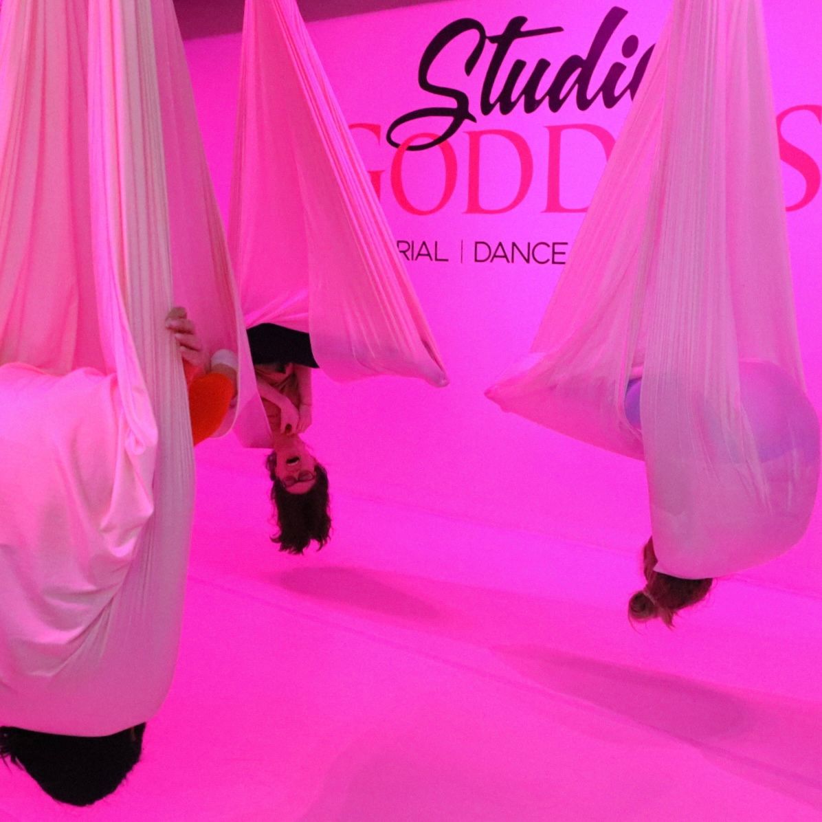 Pink-lit aerial yoga studio with a group suspended upside down in soft white fabric hammocks, playful inverted poses against a bright pink backdrop.