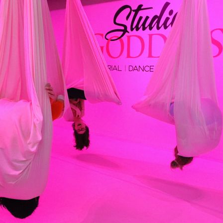 Pink-lit aerial yoga studio with a group suspended upside down in soft white fabric hammocks, playful inverted poses against a bright pink backdrop.