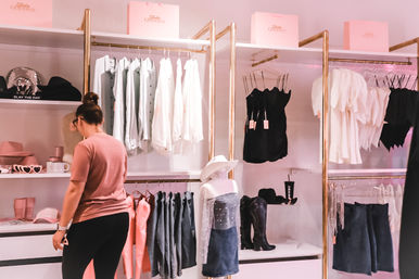 Shopper browsing a pink-themed women's boutique with gold racks showcasing white blouses, black camis, denim skirts, cowboy boots and hats, chic retail clothing display.