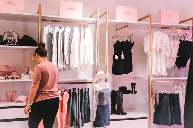 Shopper browsing a pink-themed women's boutique with gold racks showcasing white blouses, black camis, denim skirts, cowboy boots and hats, chic retail clothing display.