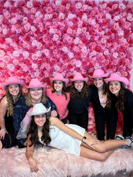 Bride-to-be in white dress and veil lounging on a faux-fur bench with five friends in pink cowboy hats in front of a pink rose wall backdrop — bachelorette party photo booth