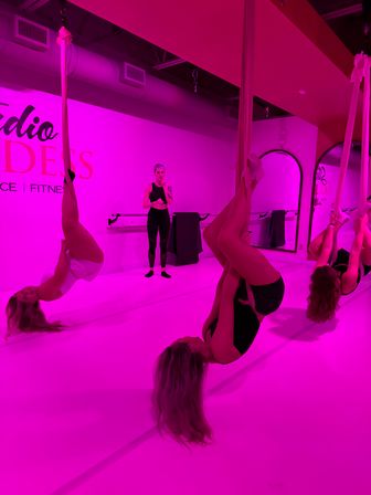 Aerial silks class in a pink-lit fitness studio: three women suspended upside down on aerial silks while an instructor watches, mirrored wall and ballet barre in the background.