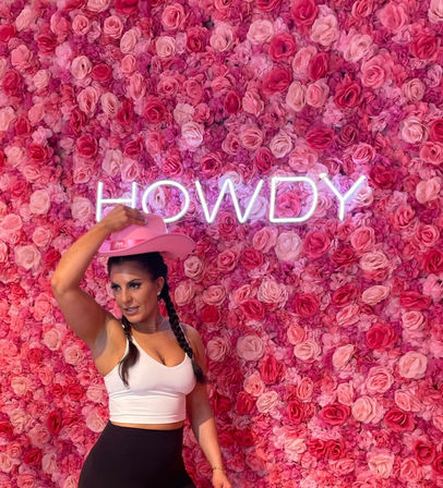 Woman in a white crop top tipping a pink cowboy hat in front of an Instagrammable pink rose wall with neon "HOWDY" sign