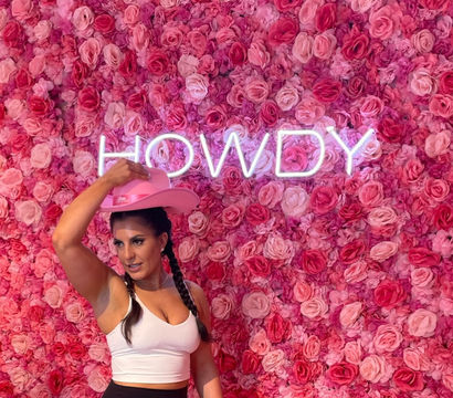Woman in a white crop top tipping a pink cowboy hat in front of an Instagrammable pink rose wall with neon "HOWDY" sign