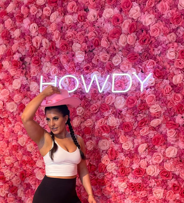 Woman in a white crop top tipping a pink cowboy hat in front of an Instagrammable pink rose wall with neon "HOWDY" sign