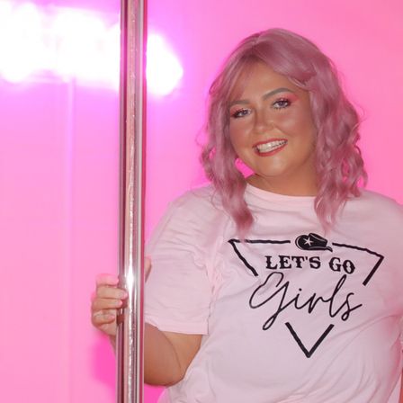 Smiling woman with pastel pink wavy hair in a bright pink backdrop, holding a chrome pole and wearing a light-pink T-shirt printed LET'S GO Girls — fun, colorful portrait.