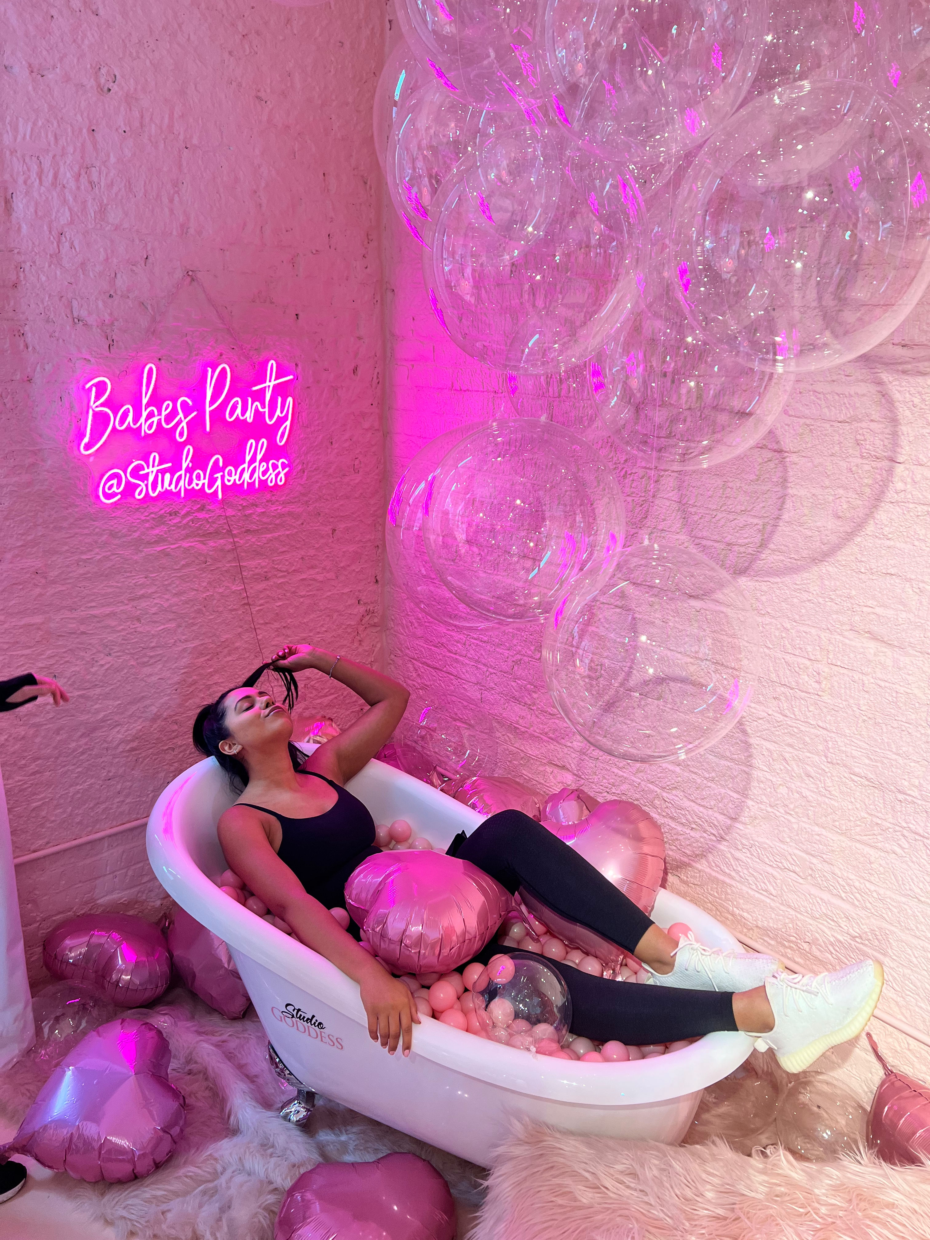 Person relaxing in a white clawfoot bathtub used as a pink ball-pit with metallic heart balloons, oversized clear bubble balloons, and a glowing pink neon party sign against a textured brick wall in a playful photo studio.
