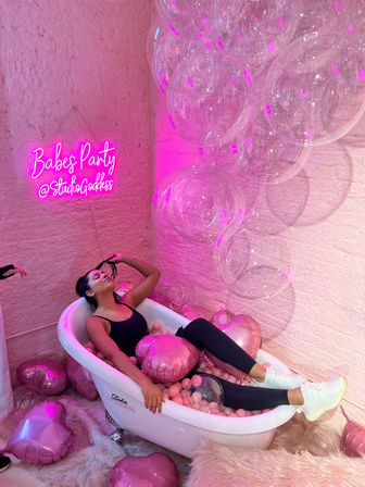 Person relaxing in a white clawfoot bathtub used as a pink ball-pit with metallic heart balloons, oversized clear bubble balloons, and a glowing pink neon party sign against a textured brick wall in a playful photo studio.
