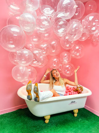 Person lounging in a white clawfoot bathtub filled with pink ball-pit balls, wearing a white tank and white boots, surrounded by clear balloons against a vibrant pink wall and green turf — playful photo studio scene.
