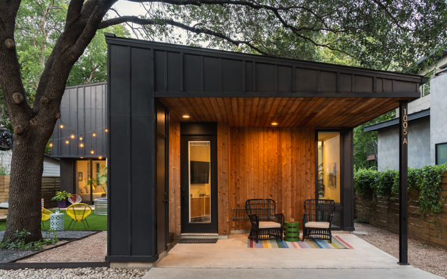 Modern tiny house with black metal exterior and warm cedar-clad covered porch, wicker chairs, colorful rug and string lights under a large oak tree