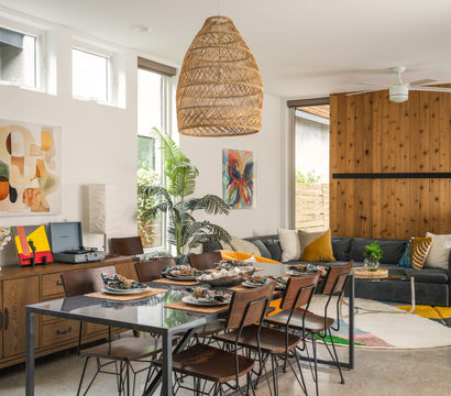 Sunlit open-plan living and dining room with wood-paneled accent wall, woven wicker pendant, dining table set for six, gray sectional sofa, indoor palm and colorful round rug.