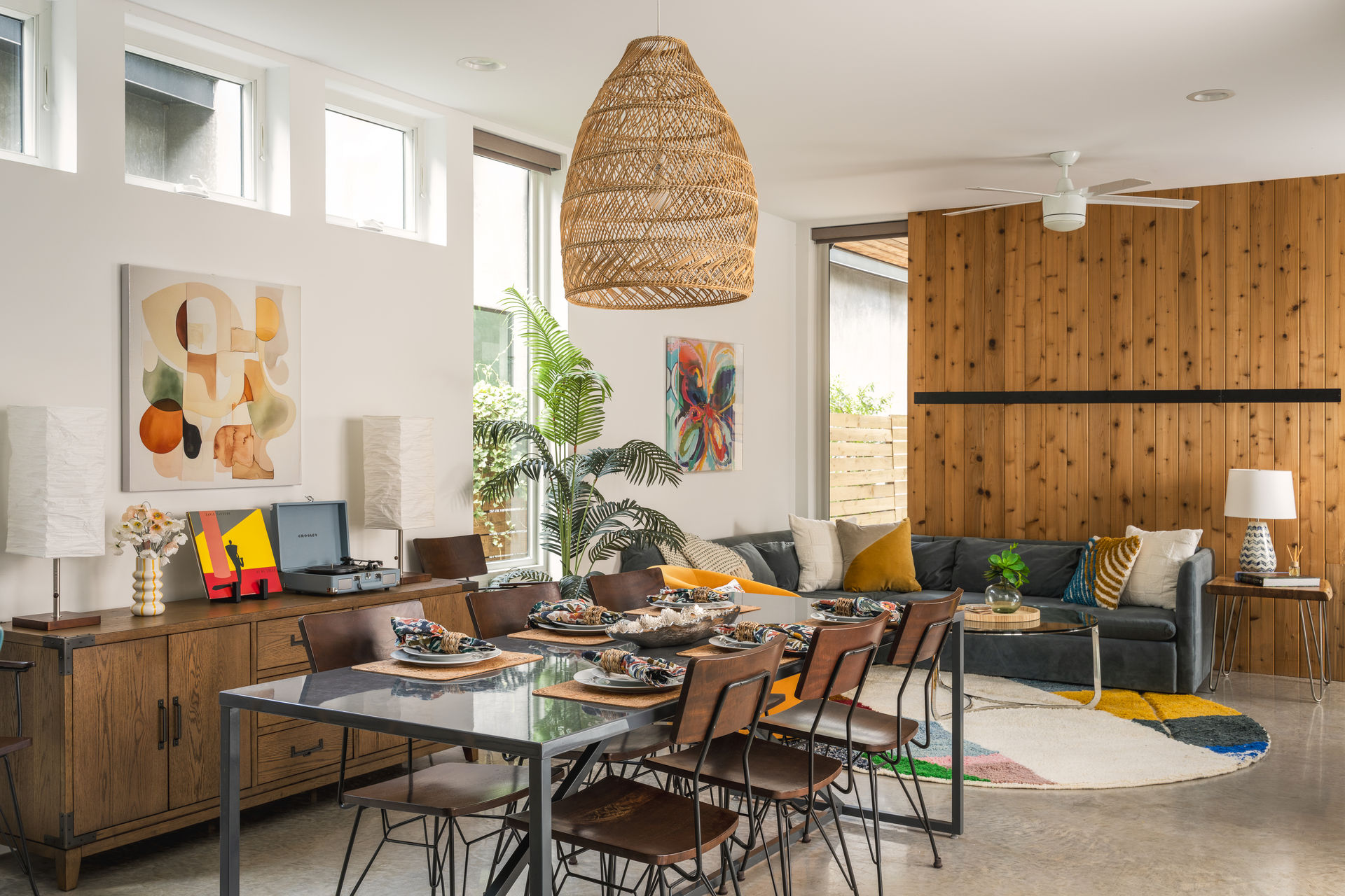 Sunlit open-plan living and dining room with wood-paneled accent wall, woven wicker pendant, dining table set for six, gray sectional sofa, indoor palm and colorful round rug.