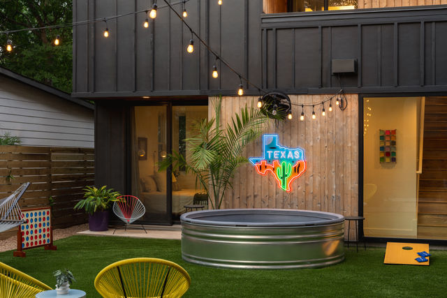 Modern Texas backyard patio at dusk with string lights, neon Texas sign featuring a cactus, round metal stock-tank pool, cornhole board and colorful outdoor chairs on artificial turf.