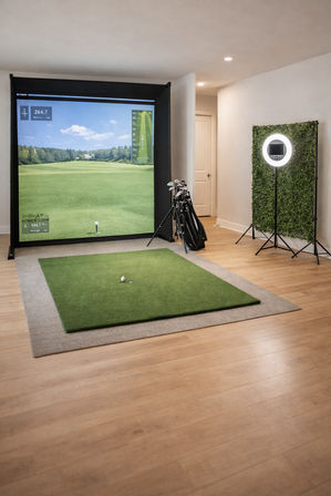 Home indoor golf simulator in a bright rec room — large projection screen showing a virtual fairway, green hitting mat with tee and ball, golf bag with clubs nearby, and a ring light with faux greenery backdrop on hardwood floor, ready to tee off.