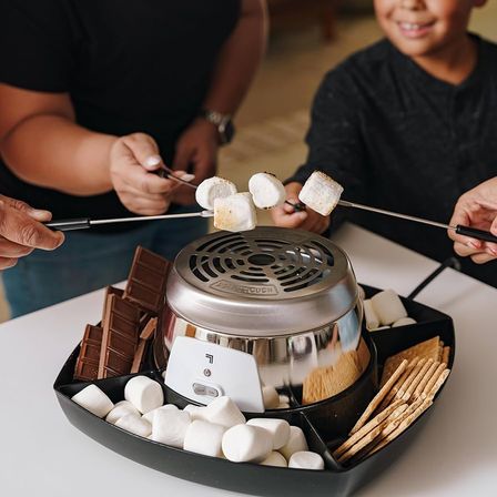 Cozy indoor s'mores moment: marshmallows on skewers toasting over a tabletop electric s'mores maker, surrounded by chocolate squares, graham crackers and extra marshmallows.