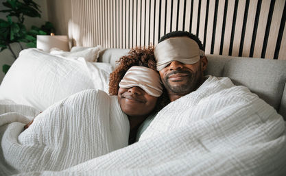 Smiling couple cuddled under a white quilt in a cozy modern bedroom, both wearing silk sleep masks and enjoying a lazy morning in bed.