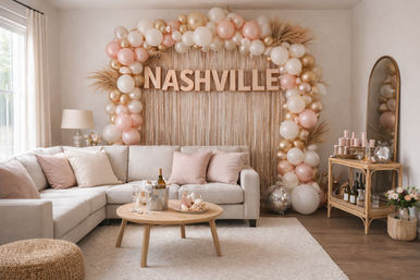 Chic Nashville-themed living room party setup with blush and gold balloon arch spelling NASHVILLE over a beige sectional, wooden coffee table with champagne and glasses, pampas grass accents and a small bar cart.