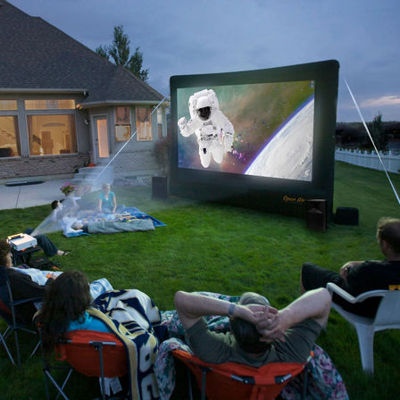 Backyard outdoor movie night at dusk with people on blankets and lawn chairs, a projector casting an astronaut-and-Earth scene onto a large inflatable screen in front of a suburban house.