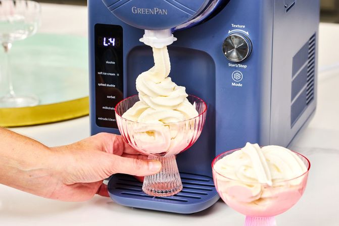 Blue countertop soft-serve machine dispensing swirled vanilla soft-serve into a pink glass dessert cup, a hand steadying the bowl with a second filled cup beside it.