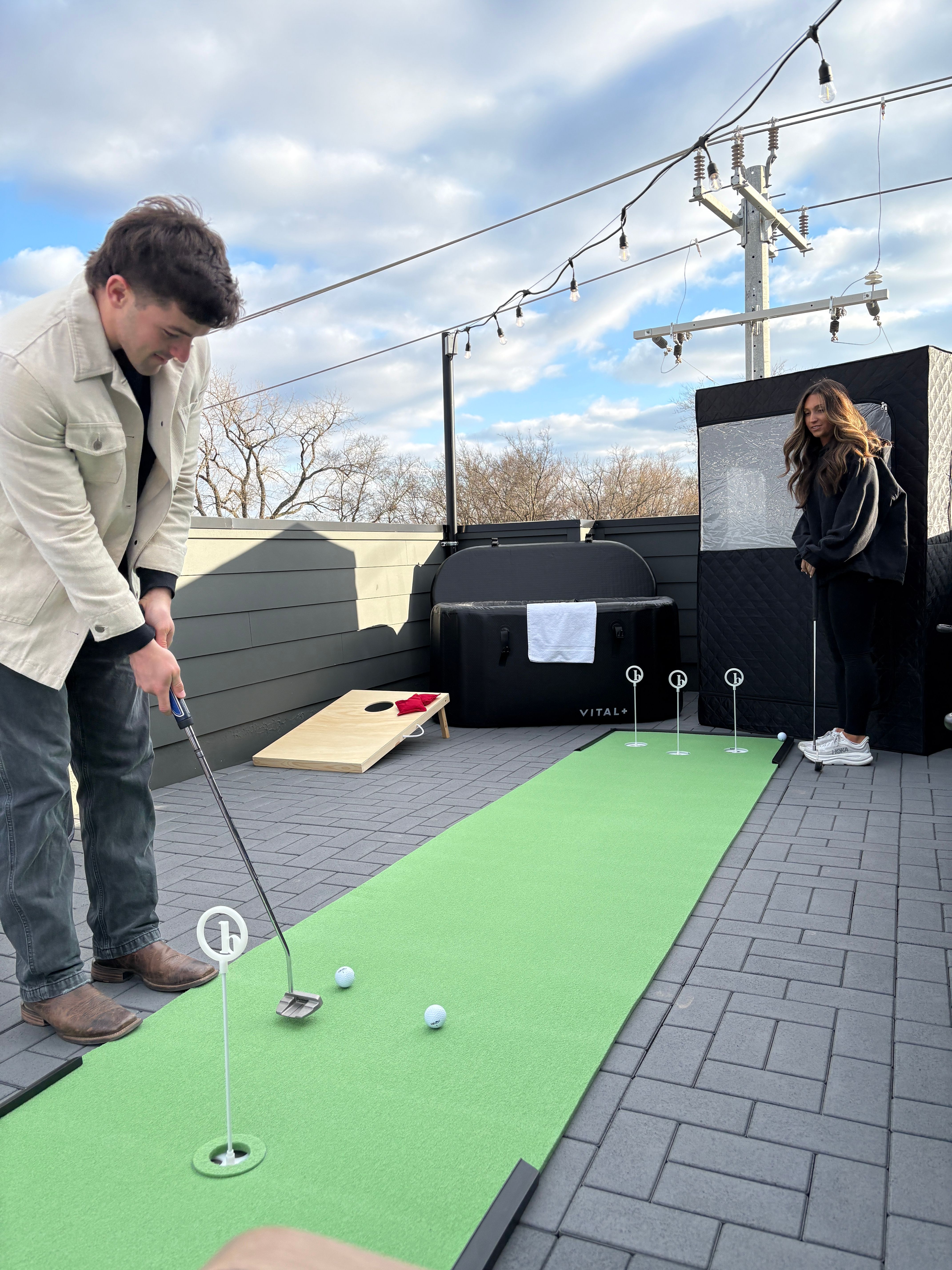 Rooftop patio scene with a portable putting green and cornhole board — person putting while another watches under string lights and a cloudy sky.