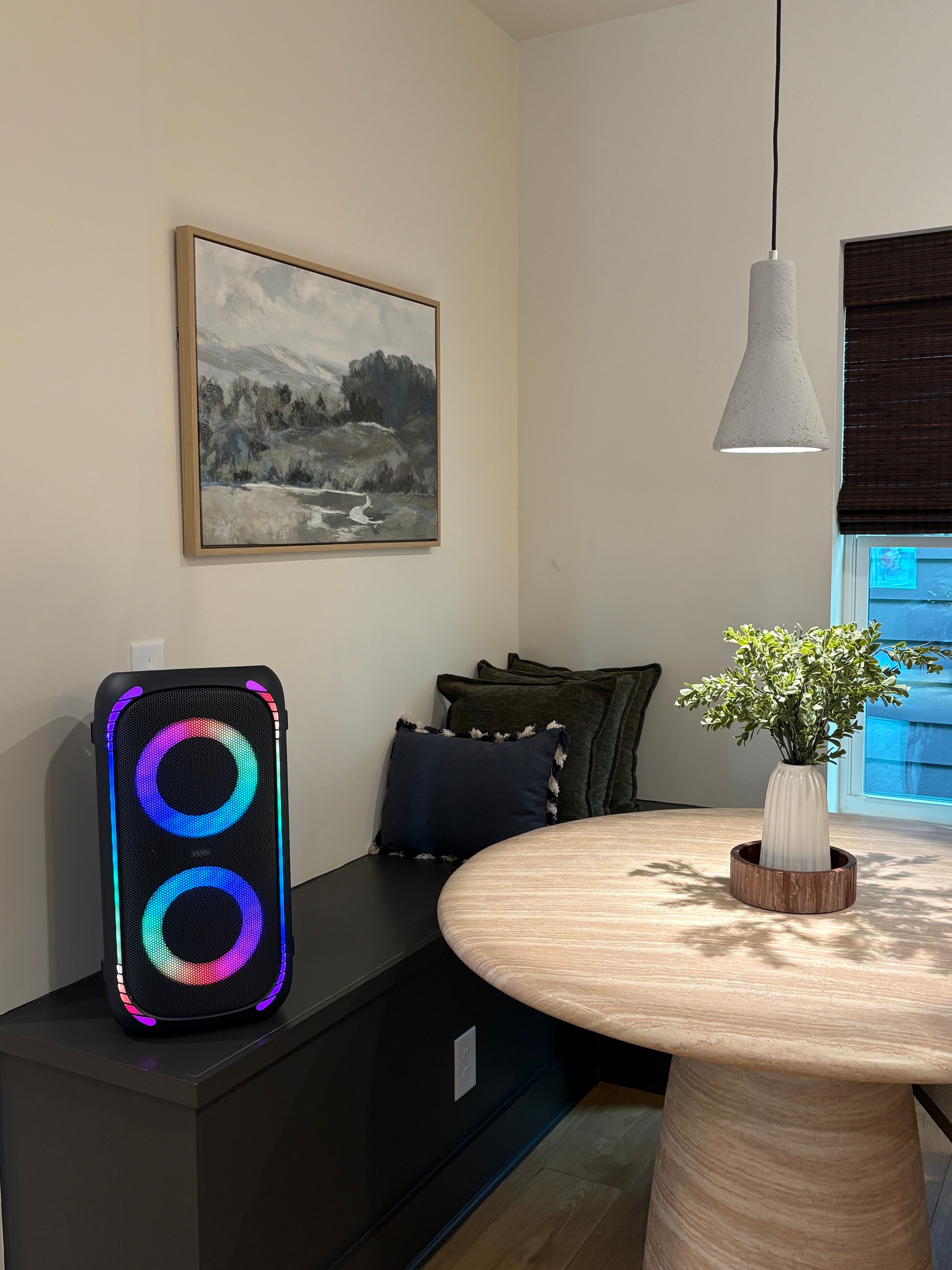 Cozy modern home dining nook with round light-wood table, white vase of greenery, hanging pendant lamp, framed landscape art, built-in bench with pillows, and a multicolored RGB LED party speaker glowing on the bench.