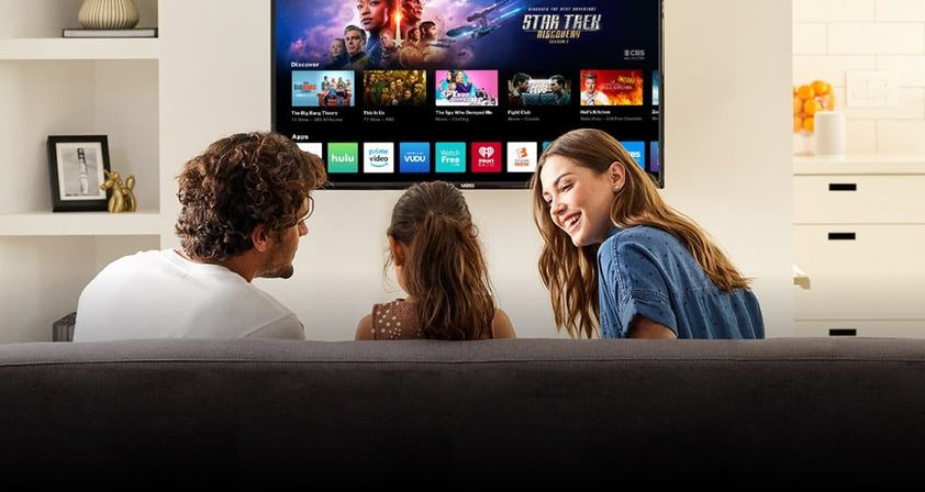Smiling family of three on a sofa in a bright modern living room, enjoying a wall-mounted smart TV streaming a space sci-fi series with streaming app icons visible.