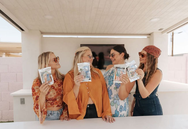 Four friends in sunglasses on a sunlit backyard patio, smiling and holding clear resealable product pouches while wearing colorful boho outfits for a casual summer moment.