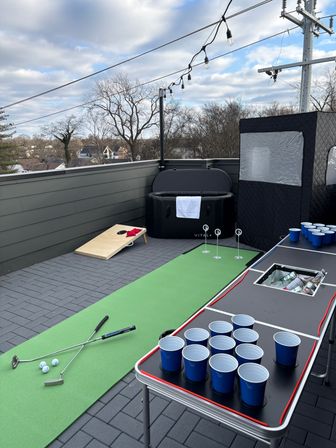 Rooftop patio with green putting mat, putters and golf balls, cornhole board, beer pong table with blue cups, inflatable hot tub, string lights and suburban tree-lined view under a cloudy sky.