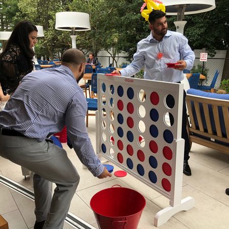 People playing a giant Connect Four on an outdoor patio event — red and blue discs and bucket, one player wearing a balloon crown