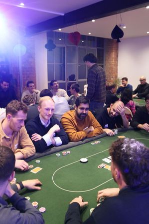 Group playing poker at a green-felt table during a casual poker night in a loft-style room with exposed brick, hanging card-suit decorations, and stacks of chips and cards on the table.