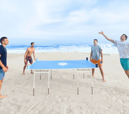 Four friends playing a blue portable table game with cups on a sandy ocean beach, waves rolling in under a bright sky