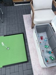 Backyard patio with cushioned wooden lounge chair, open white cooler of ice-cold sodas and sports drinks, and a small green putting mat on gray paver tiles