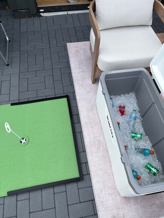 Backyard patio with cushioned wooden lounge chair, open white cooler of ice-cold sodas and sports drinks, and a small green putting mat on gray paver tiles