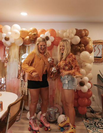 Two women roller-skating at an indoor retro disco party in a home — warm-toned balloon arch, fringe backdrop, mirrored disco balls and smiling celebration.