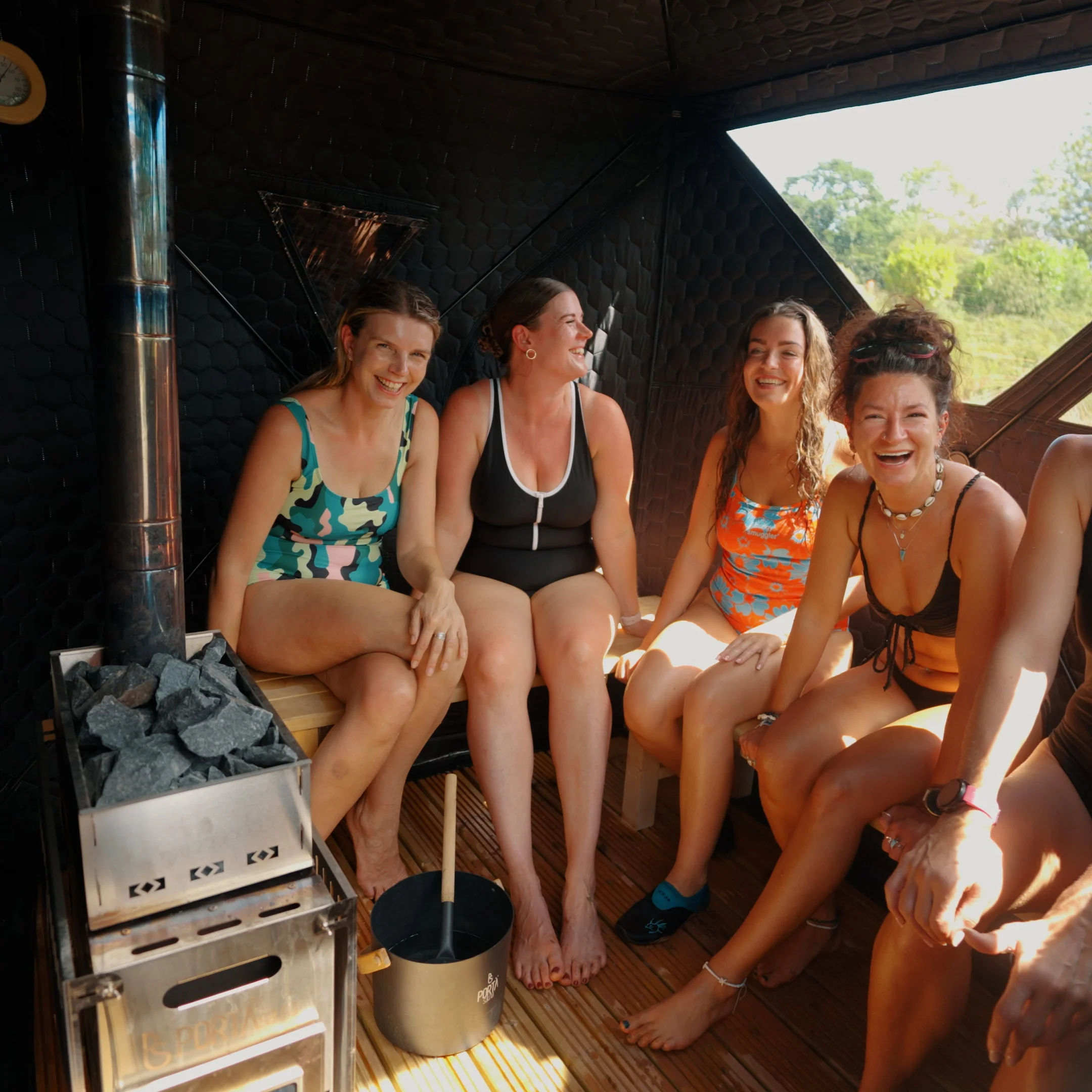 Four women in swimsuits laughing together in a sunlit wooden outdoor sauna beside a hot-stone stove with a green forest view